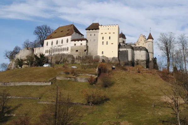 Das mittelalterliche Schloss Lenzburg thront auf einem steilen, grastragenden Felsberg vor leicht bewölktem blauem Himmel. Historische Mauern, Türme und Gebäude prägen die Anlage.