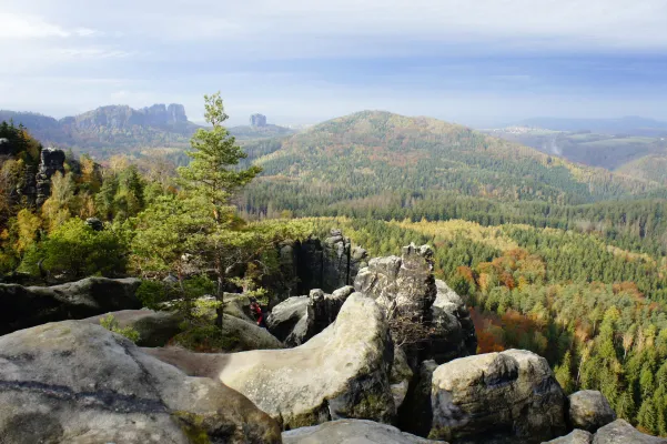 Panoramablick von einer Felsterrasse über weitläufige Herbstwälder und eindrucksvolle Sandsteinklippen unter leicht bewölktem Himmel.