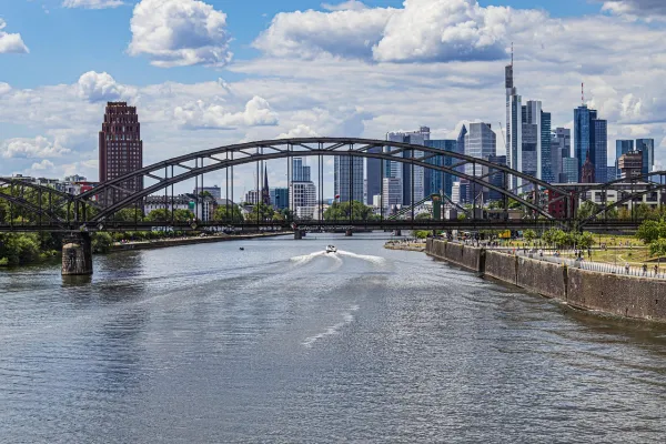 Blick über den Main auf eine Bogenbrücke mit einem lokalen Boot; im Hintergrund die Frankfurter Skyline unter wolkigem Himmel.