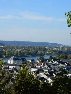 Aussicht von oben auf die Dächer der Altstadt von Königswinter. Im Hintergrund fließt der breit geschwungene Rhein, dahinter erstreckt sich die grün bewaldete Hügellandschaft des gegenüberliegenden Ufers unter blauem Himmel.